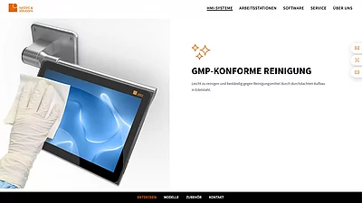 Hand mit Einweghandschuh reinigt einen industriellen Touchscreen aus Edelstahl, montiert an einer Wandhalterung, dargestellt auf einer Website mit dem Hinweis „GMP-konforme Reinigung“.